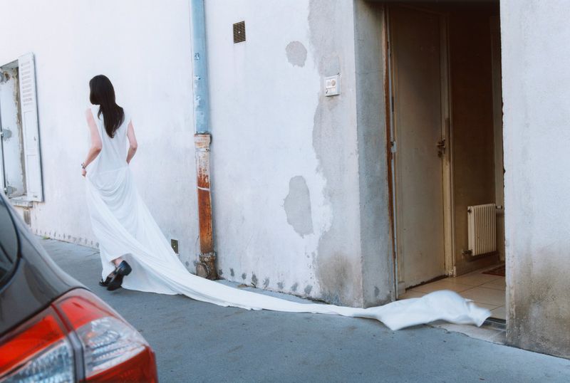 Woman in white dress walking on an outdoor walkway next to buildings, with a car tail light visible in the foreground.