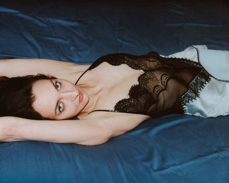 Brunette woman in black and white lace lingerie lying on a blue satin bedsheet, looking at the camera with direct gaze.