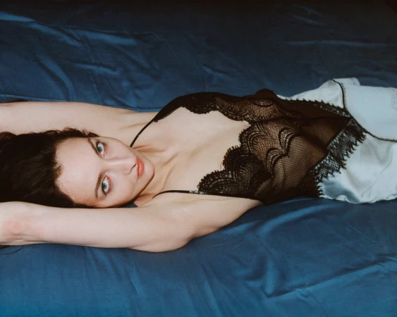 Brunette woman in black and white lace lingerie lying on a blue satin bedsheet, looking at the camera with direct gaze.