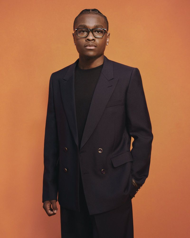 Young Black man wearing a dark double-breasted blazer and glasses against a warm solid orange background