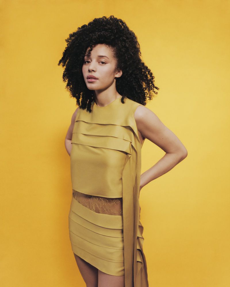 Portrait of a woman with curly hair in a short yellow dress, posing against a bright yellow backdrop in a studio setting.