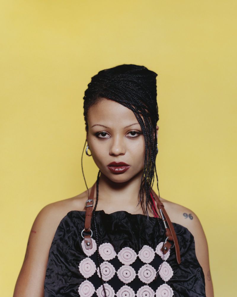 Portrait of a young woman with braided hair against a yellow backdrop, wearing a dark top with light-colored floral crochet embellishments.