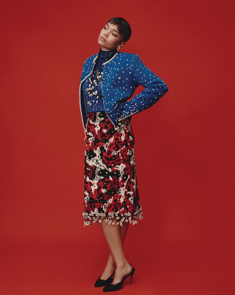 A woman wearing a blue patterned jacket and red floral skirt posing against a solid red studio background.