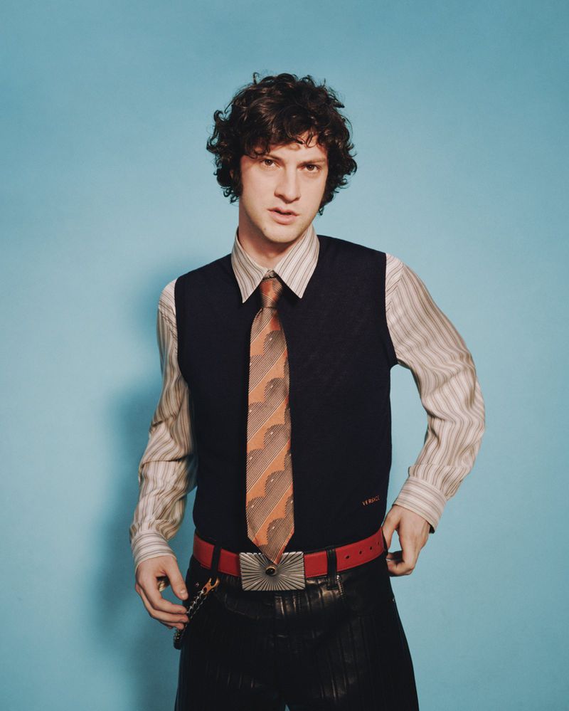 A stylish adult male in a vest and tie poses against a blue backdrop. He has curly hair and is wearing a red belt with a large buckle.