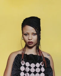 Portrait of PinkPantheress against a yellow background wearing a black top with crocheted circles and brown straps.