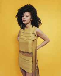 Young woman with dark curly hair wearing a gold layered dress against a matching yellow backdrop.