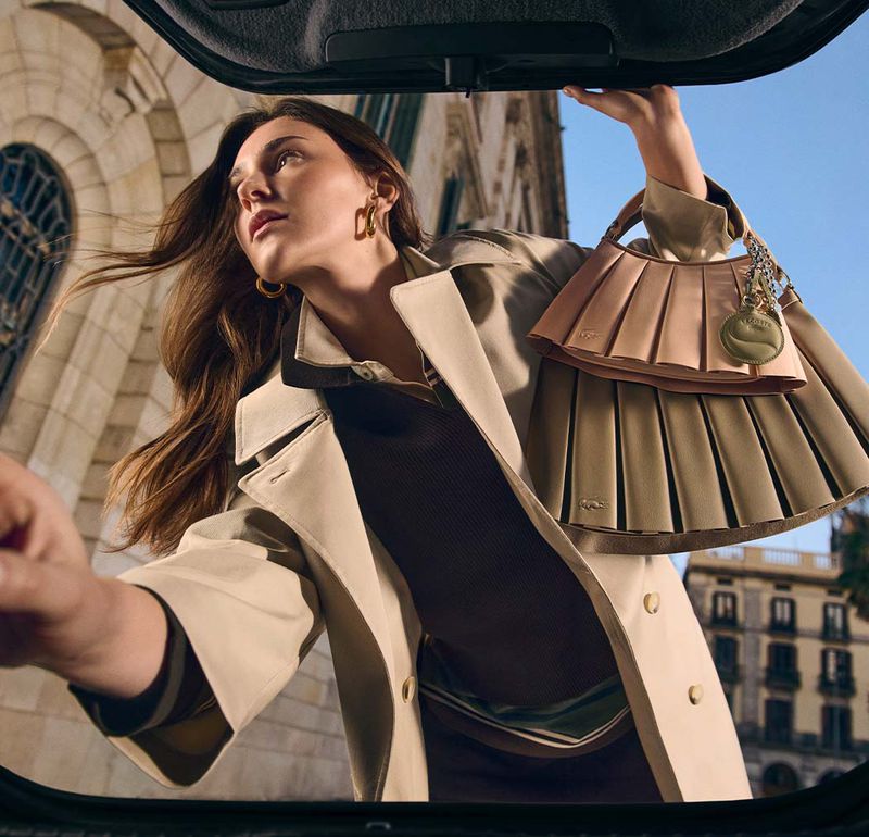 Elegant woman in a trench coat loading a stylish pleated handbag into the back of a car in front of an old building.