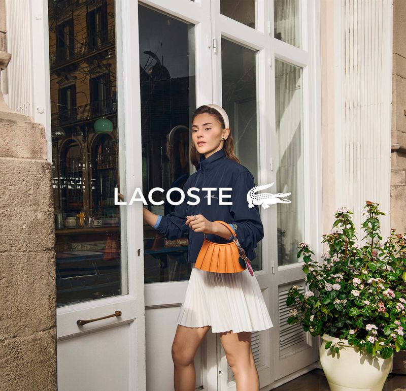 A woman in a navy jacket, white pleated skirt, and headband exits a white door with a Lacoste logo next to her head.