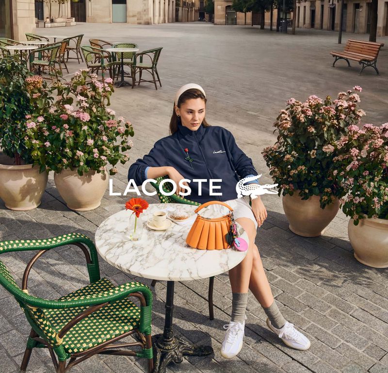 A woman sits at a table on a city street, with a Lacoste purse and flower, beside plants. The Lacoste logo is displayed.