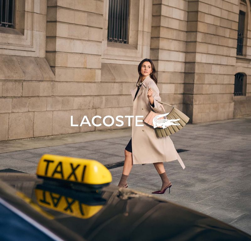 A woman wearing a trench coat walks on a city street carrying a Lacoste handbag, with a yellow taxi cab in the foreground.