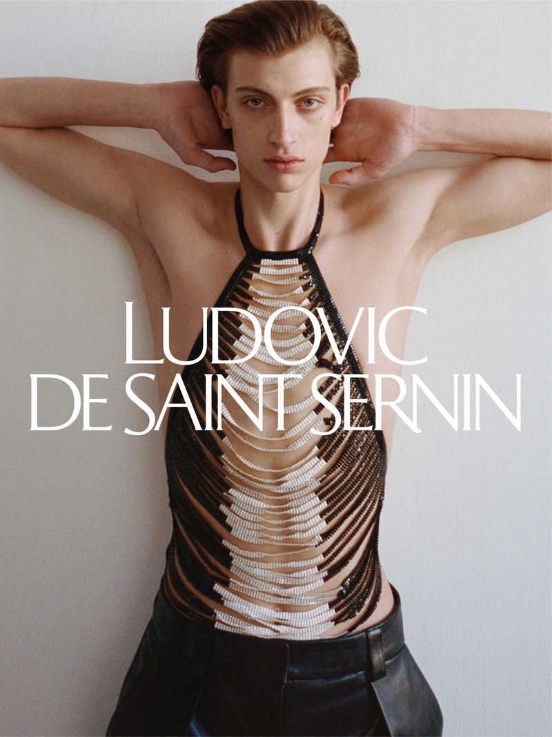 A fair-skinned male fashion model poses in a designer beaded halter top and leather pants against a neutral studio backdrop.