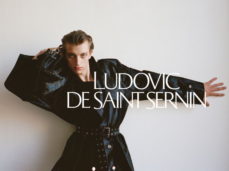 Young man poses against white wall wearing black, belted coat and fashionable attire. Fashion photography for Ludovic de Saint Sernin.