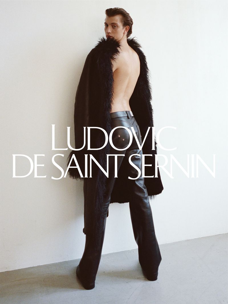 Man with bare back, wearing an open black fur coat and black leather trousers, stands in a photo studio in a fashion pose.
