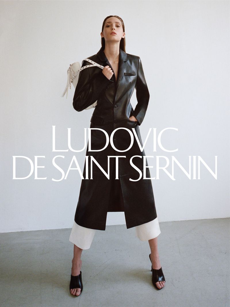 A woman poses in a black coat with a white bag against a white background, wearing white pants and black heels. Fashion photography.