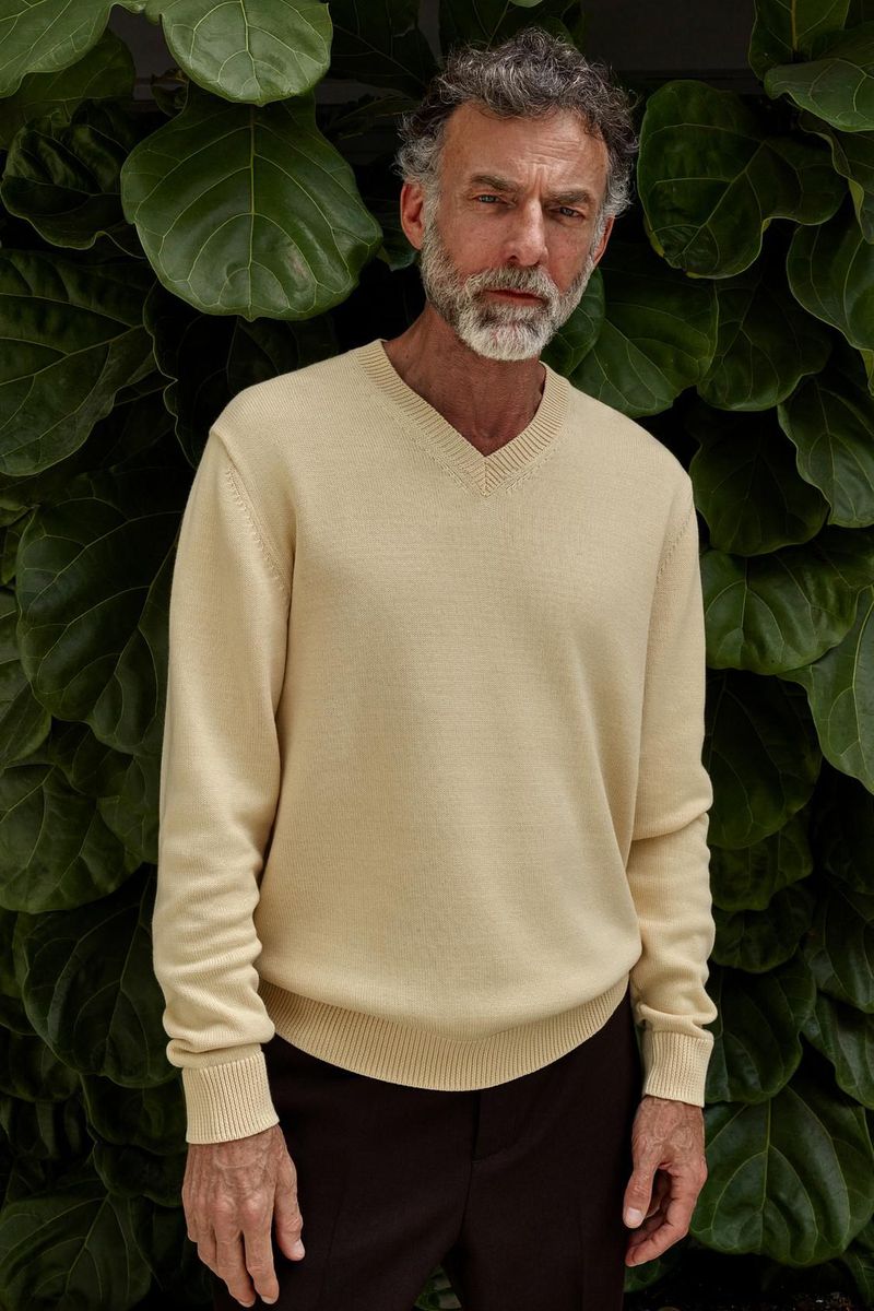 Middle-aged man with grey hair and beard poses in a cream-colored V-neck sweater against a backdrop of large green leaves.