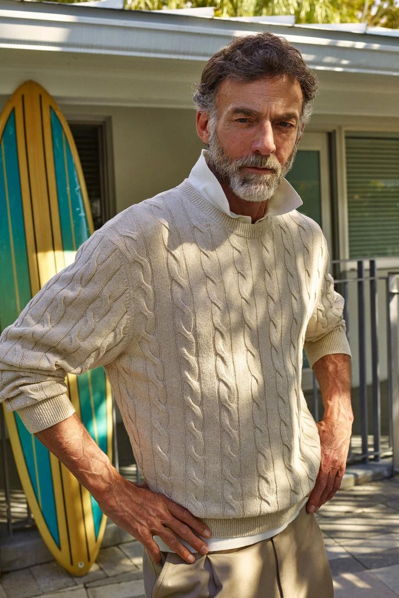 Mature man with salt-and-pepper hair and beard poses in a cream cable-knit sweater, with a surfboard in the background.
