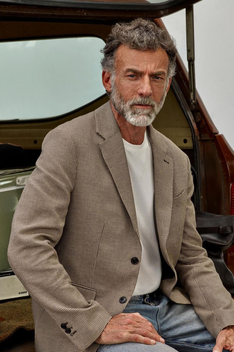 Mature man seated near an open car trunk, wearing a patterned blazer, white tee, and jeans, looking directly at the camera.
