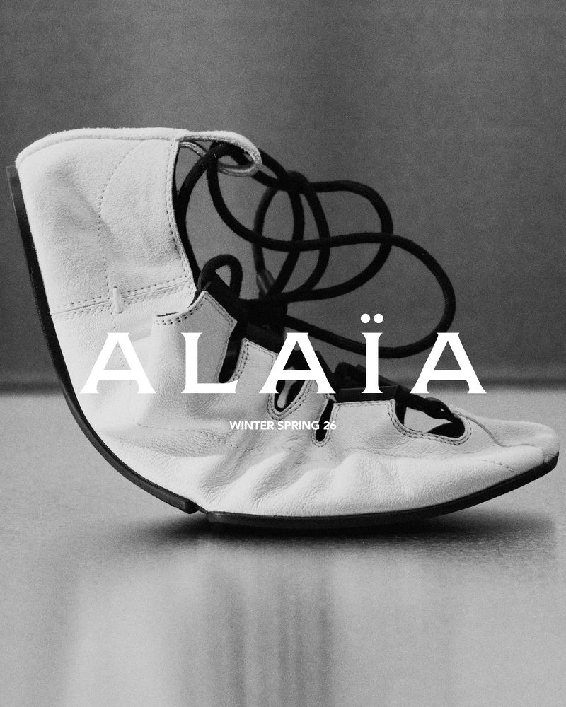 Alaïa sandal in a studio shot. The cream-colored flat sandal has black laces and is photographed against a gray backdrop. Fashion.