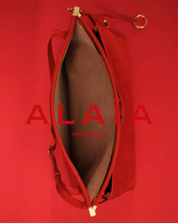 Red leather shoulder bag with gold hardware and beige lining on a matching red backdrop, featuring the ALAÏA Winter Spring logo.