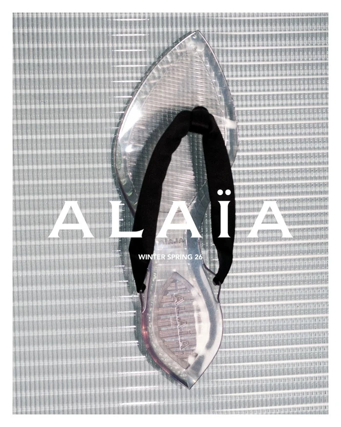Clear transparent sandal with a black thong strap positioned vertically against a textured grey linear background.