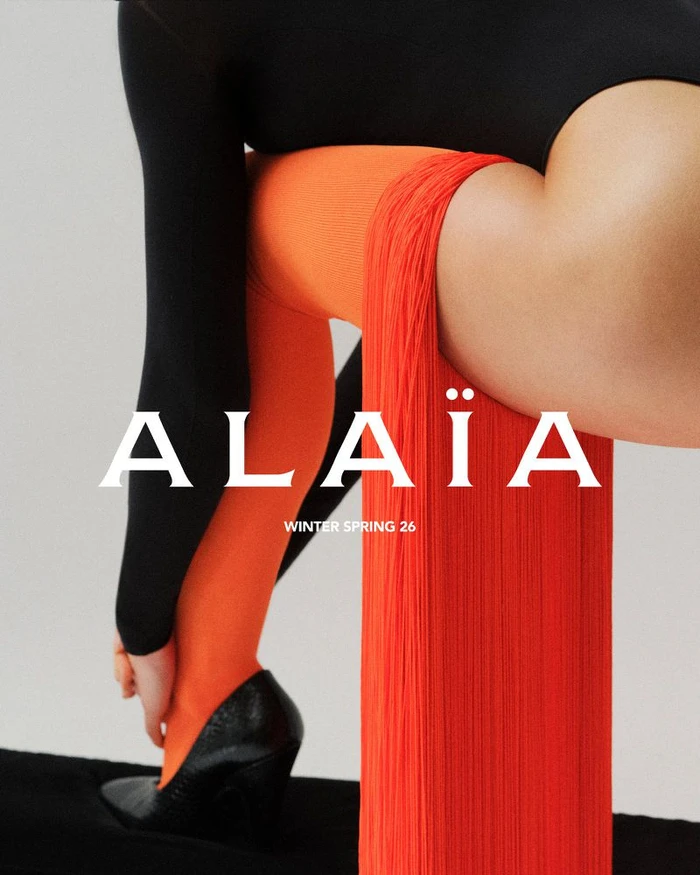 Model wearing black top, orange tights, and black shoes seated on a bright red fringed column against a light background