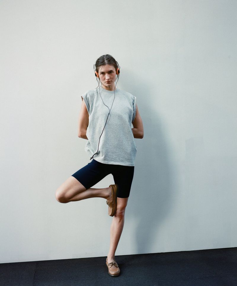 Woman in grey top and navy shorts balances in tree pose against a white wall, wearing brown loafers and black headphones.