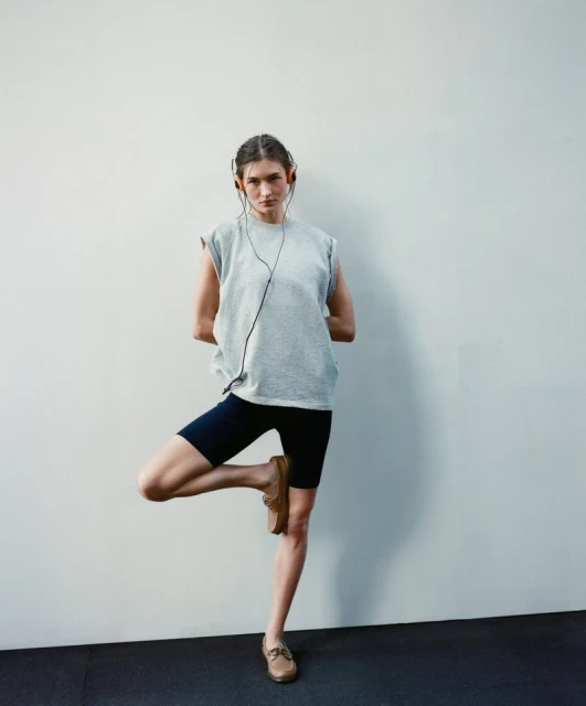 Woman in grey top and navy shorts balances in tree pose against a white wall, wearing brown loafers and black headphones.