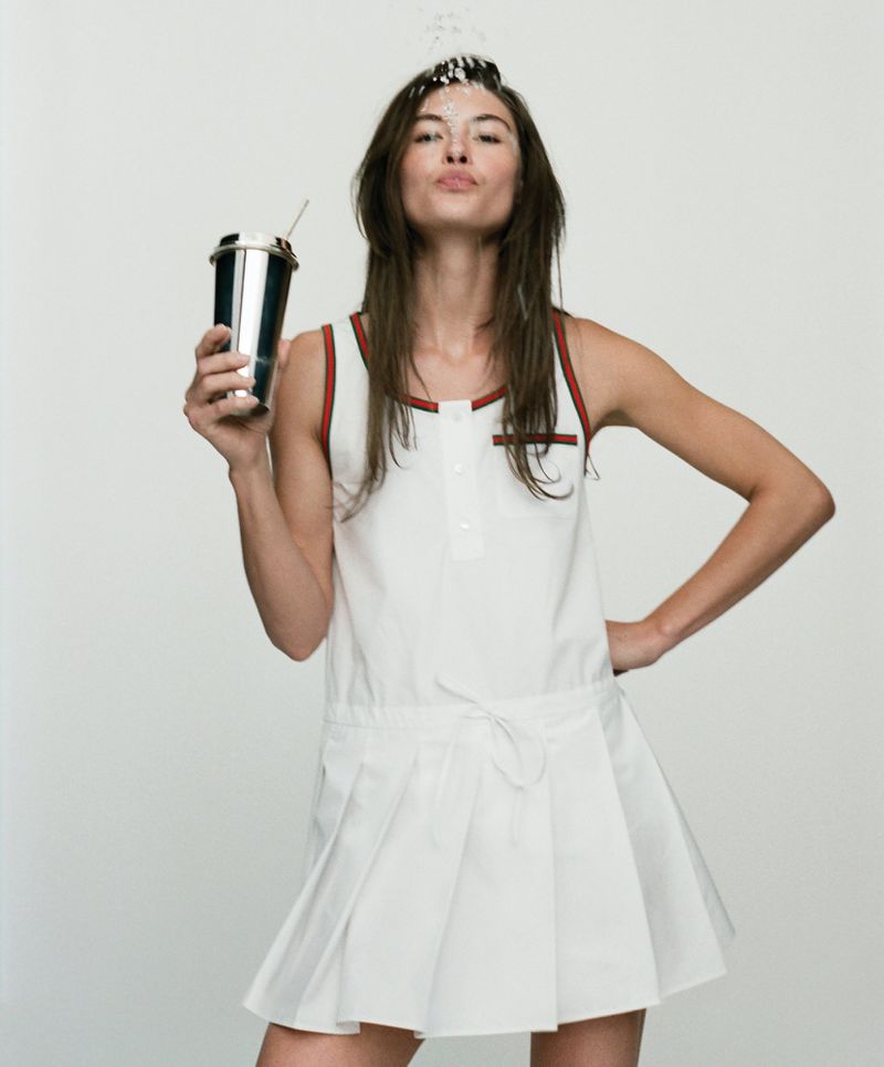 Adult woman in a tennis dress pours water over her head, holding a stainless steel cup with a straw in a studio shot.