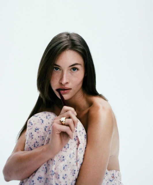 Close-up of a young adult female model with long brown hair posing with a pen between her lips in a light and bright studio setting.