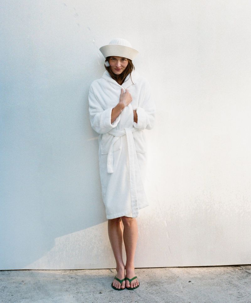 Woman in white robe and sailor hat standing against a white wall, wearing green flip-flops, full shot, bright lighting, indoor setting.