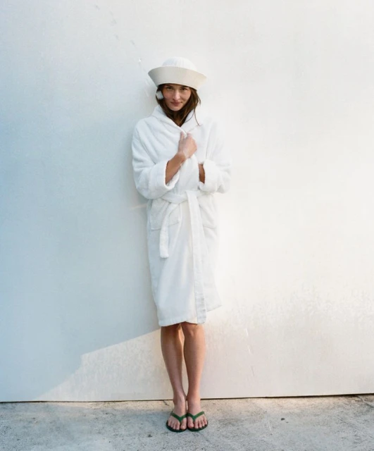 Woman in white robe and sailor hat standing against a white wall, wearing green flip-flops, full shot, bright lighting, indoor setting.