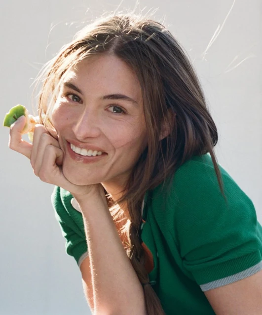 Portrait of a woman with braided hair smiling and holding a piece of fruit in her hand against a light background.