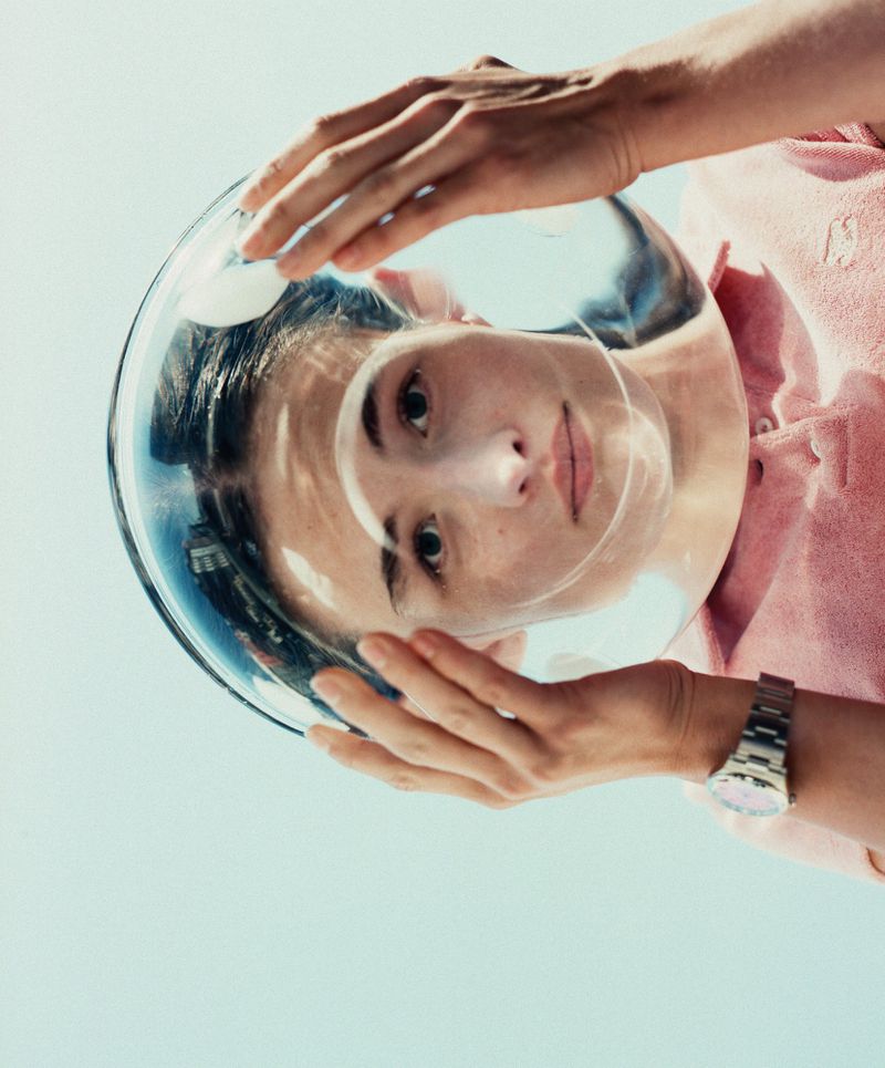 Woman with clear space helmet, hands on the edges, pink top, silver watch, looks up at the camera against blue sky, eye-level.