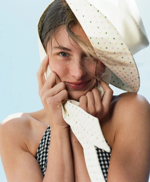 Close-up portrait of a smiling woman wearing a patterned white bonnet, with a light blue background, capturing a soft and natural aesthetic.