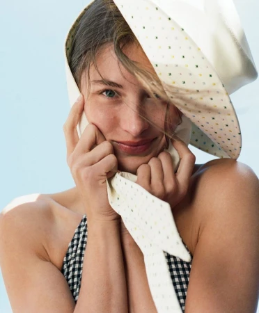 Close-up portrait of a smiling woman wearing a patterned white bonnet, with a light blue background, capturing a soft and natural aesthetic.