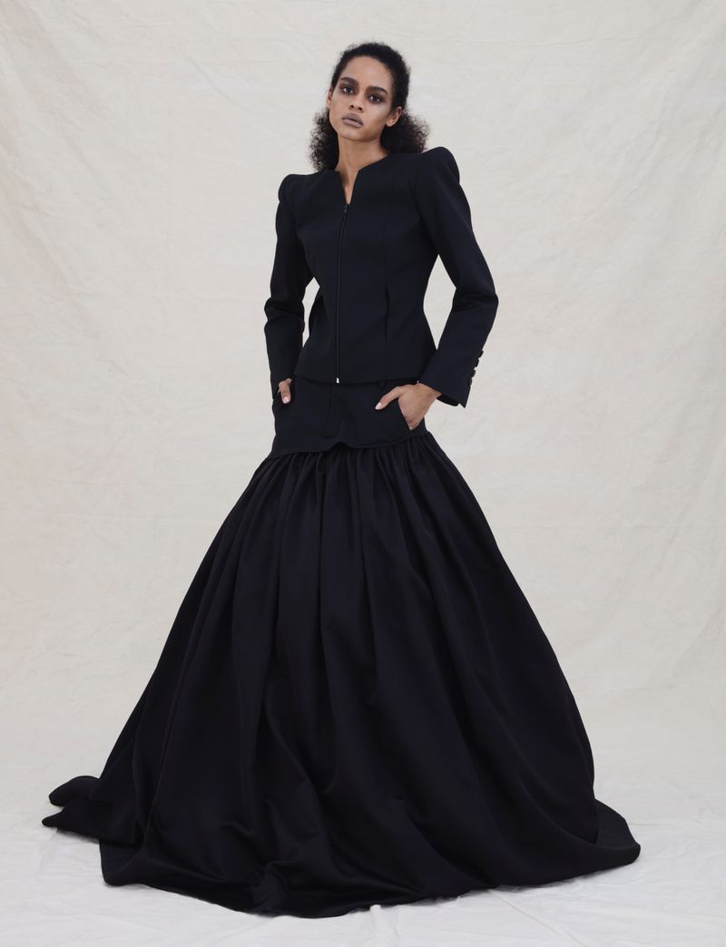 Model wearing a black structured jacket and voluminous full skirt standing against a neutral textured studio backdrop.