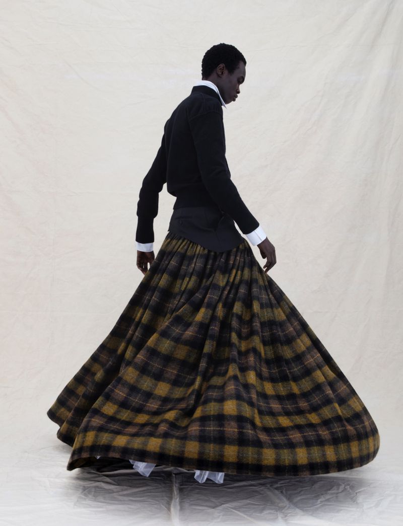 Black model in a plaid skirt and black top walking in a studio setting against a cream backdrop, showcasing fashion and style.
