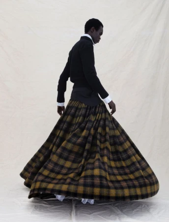 Black model in a plaid skirt and black top walking in a studio setting against a cream backdrop, showcasing fashion and style.