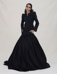 Model wearing a black structured jacket and voluminous full skirt standing against a neutral textured studio backdrop.