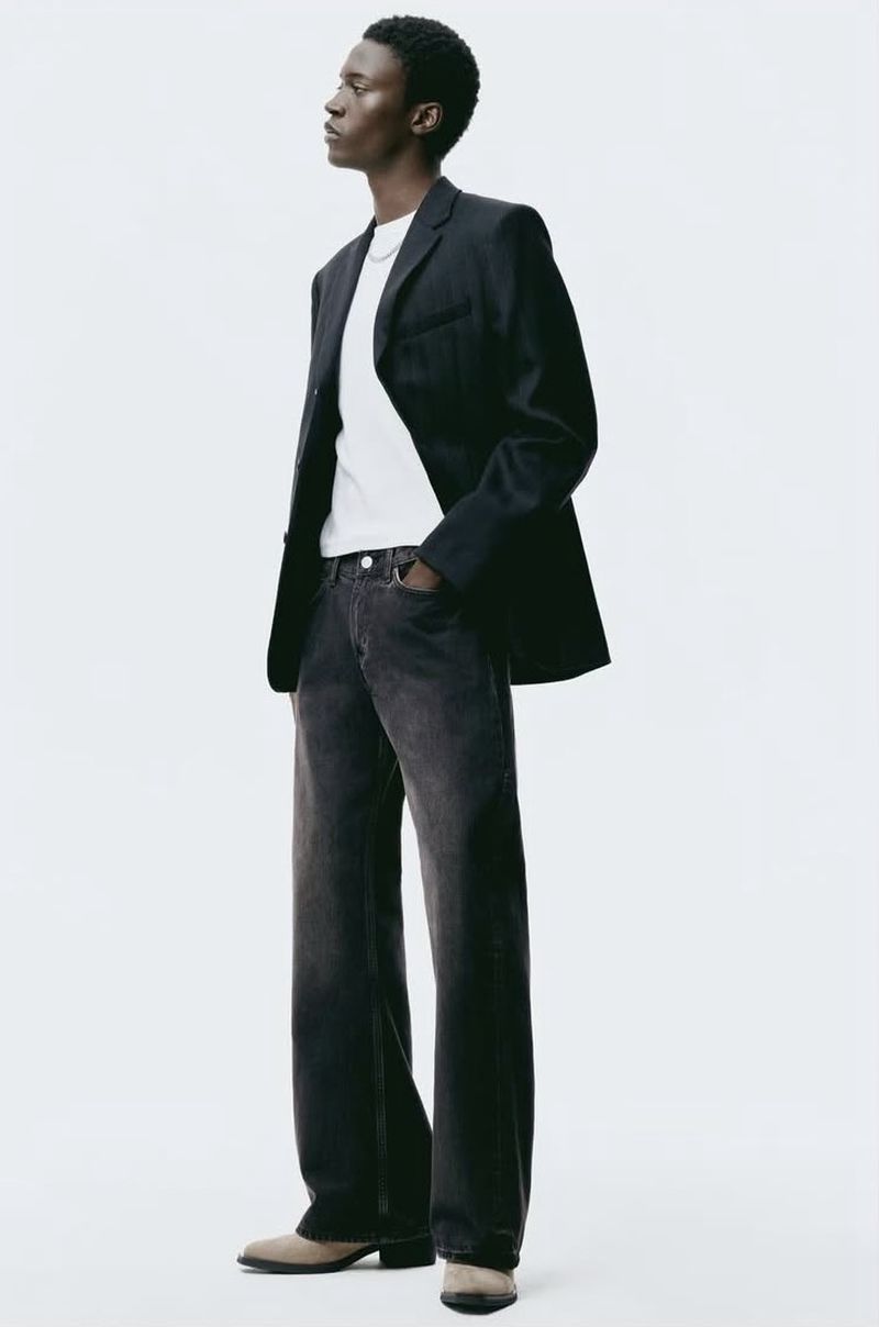 Man in dark jeans and blazer, standing in a full shot against a white backdrop, fashion pose, dark hair, in studio lighting.