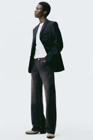 Man in dark jeans and blazer, standing in a full shot against a white backdrop, fashion pose, dark hair, in studio lighting.