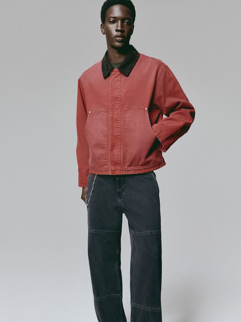 A young adult male poses in a coral jacket and dark wash jeans against a neutral backdrop in a full shot.