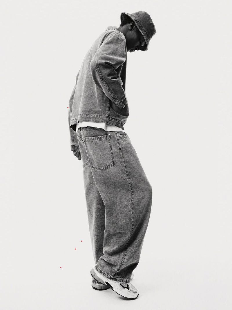 Monochrome fashion shot of a Black male model wearing a denim jacket, loose jeans, a bucket hat, and sneakers on a white background.