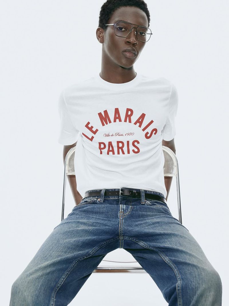 Man with dark hair, wearing glasses, t-shirt with Le Marais Paris, and jeans sits on a transparent chair against a white backdrop.
