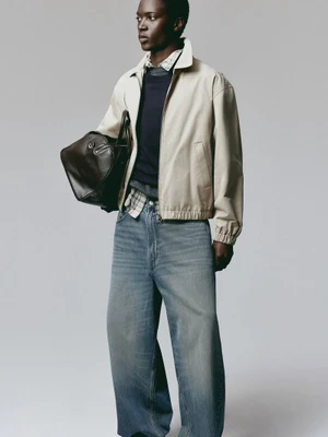 Model wearing a light beige jacket, dark sweater, and loose blue jeans holding a dark leather bag against a grey background.