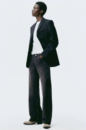 Model wearing a black blazer, white t-shirt, and dark denim jeans standing against a plain white background