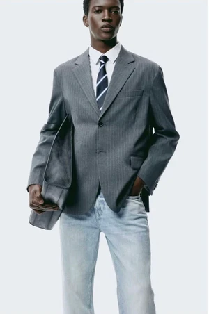 Black man in a grey pinstripe blazer, white shirt, striped tie, and light blue jeans holding a large bag against a white backdrop.