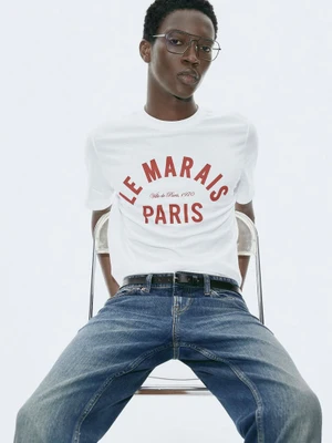 Model wearing a white graphic t-shirt and blue denim jeans seated against a clean white backdrop