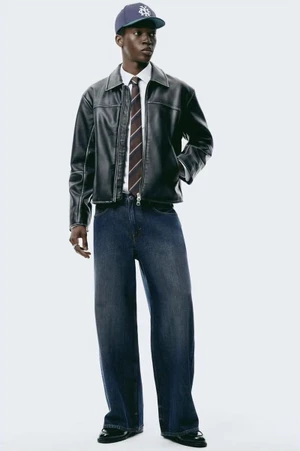 Model Limamu Mbaye wearing a black leather jacket, striped tie, and denim jeans against a plain white studio background.