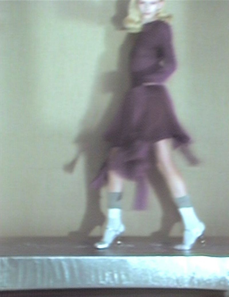Woman in purple dress and white socks poses in front of a muted wall, casting a shadow, in a fashion or editorial style shot.